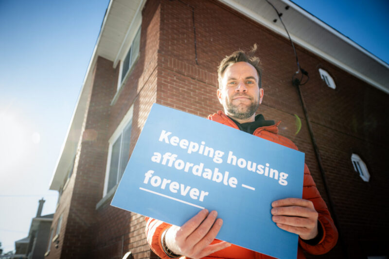 Un homme vêtu d'une veste orange tient une pancarte sur laquelle on peut lire « Pour que le logement reste abordable — pour toujours » devant une maison en briques.