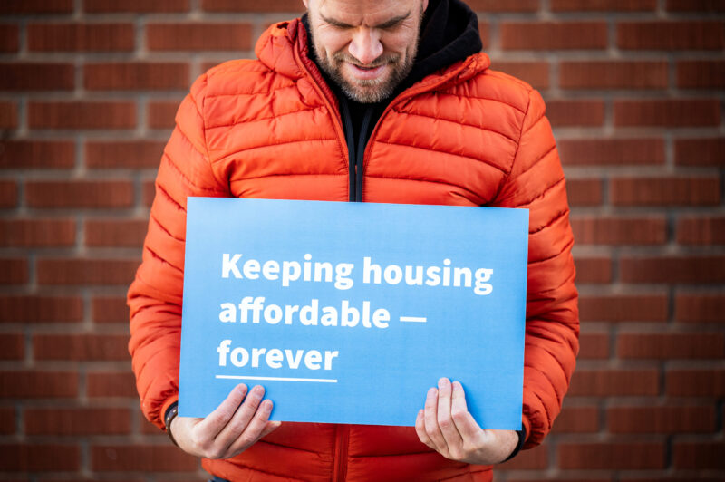 Person in orange jacket holds blue sign that says, Keeping housing affordable — forever, against brick wall.