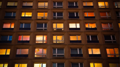 An affordable housing apartment facade at dusk, with warmly lit windows and some dark, unlit ones.