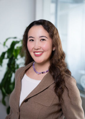 Smiling woman with wavy hair in a brown blazer and purple necklace, standing indoors near a plant.