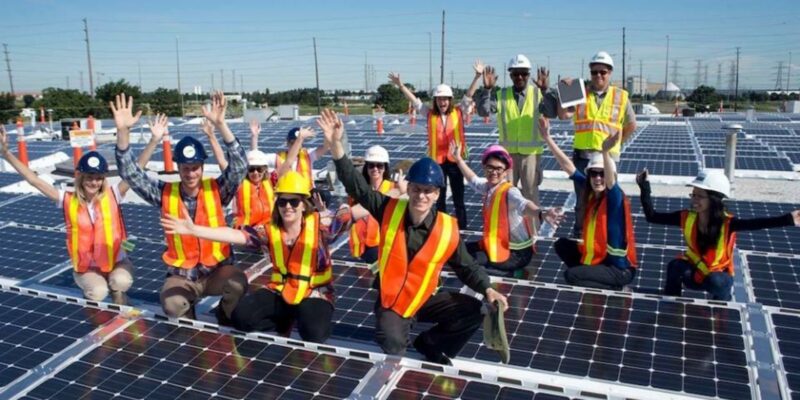Un groupe de personnes portant un équipement de sécurité, souriantes et levant les mains, sur un chantier d'installation de panneaux solaires.