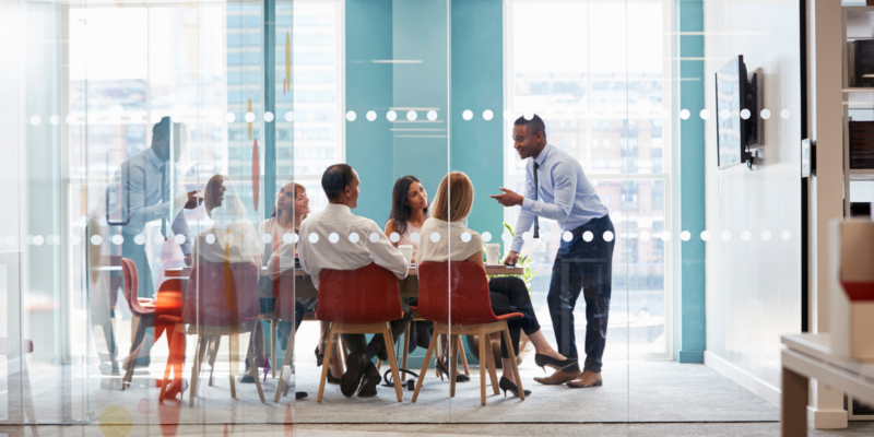A man presents to a group seated in a modern glass-walled meeting room, weaving ideas into a tapestry of insight.