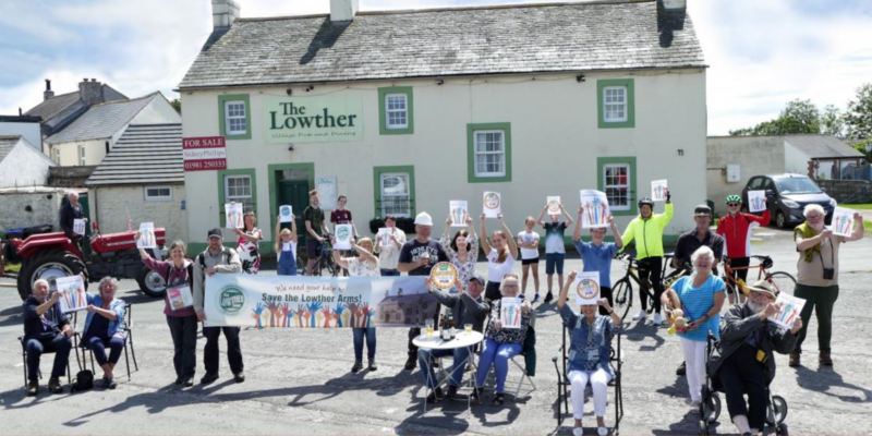 A group protests with signs at The Lowther pub, highlighting UK social investment amid its For Sale sign.