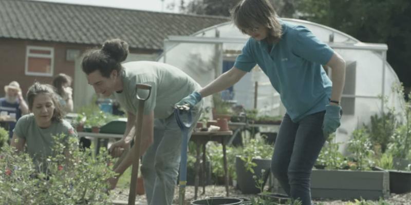 Trois personnes jardinent ensemble en plein air près d'une serre, souriantes, tout en discutant d'investissement à impact social avec les plantes.