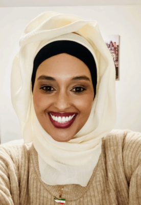Woman in a cream hijab and beige top smiling in a well-lit room with framed art on the wall.