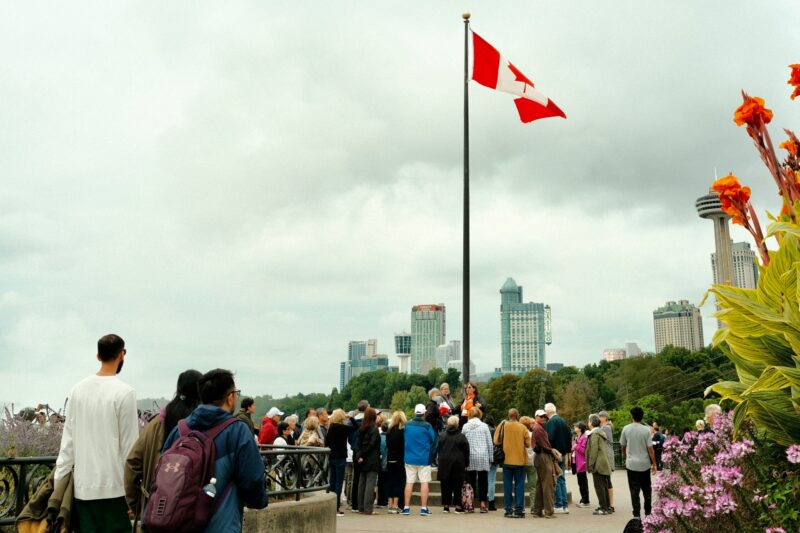A group of people gather near a Canadian flag with city buildings and flowers in the background.