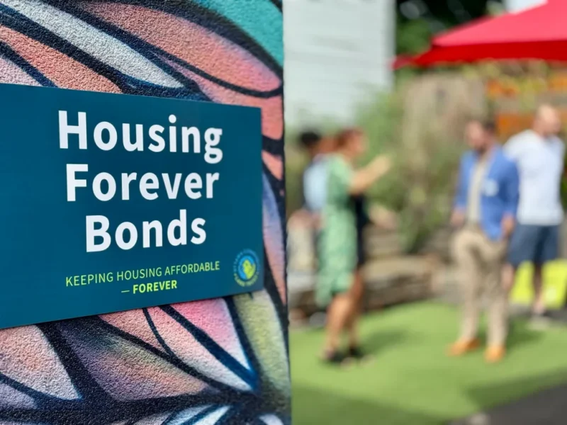 Une affiche portant l'inscription « Housing Forever Bonds » est visible, avec des personnes en train de discuter dans un décor en plein air, sur un arrière-plan flou.