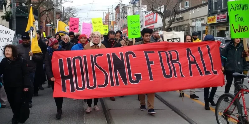 Les manifestants défilent en brandissant une grande banderole rouge sur laquelle on peut lire « UN LOGEMENT POUR TOUS », afin de sensibiliser le public au problème de l'offre de logements.