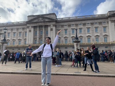Smiling person poses with arms raised in front of Buckingham Palace; crowds and blue sky in the background.