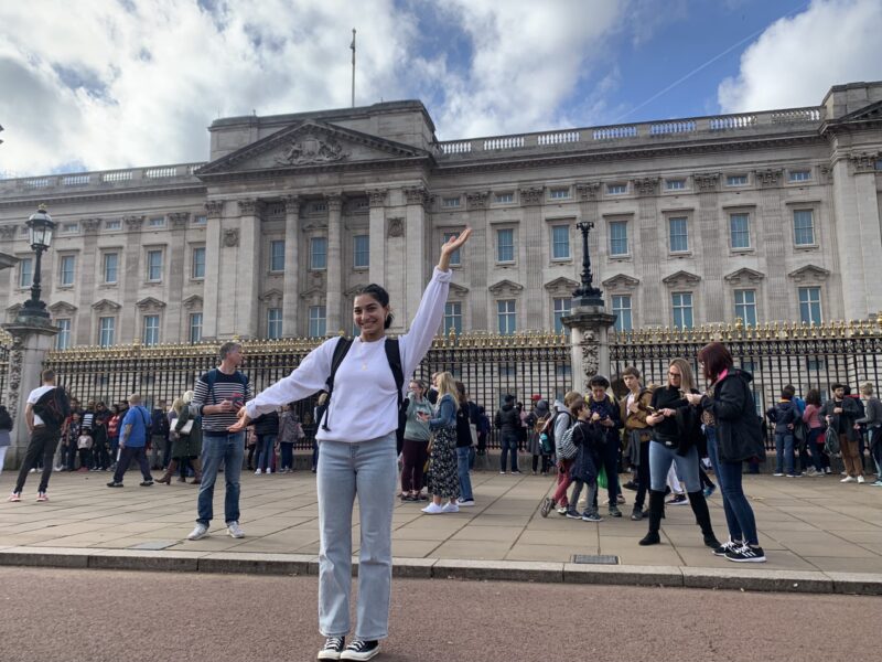 Smiling person poses with arms raised in front of Buckingham Palace; crowds and blue sky in the background.
