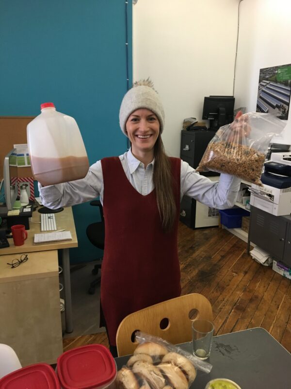 Woman in a knit hat holds a jug of cider and a bag of granola, smiling in an office kitchen.