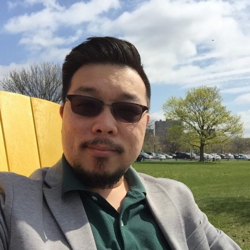 Man in sunglasses sitting on a yellow chair outdoors with trees, cars, and buildings in the background.