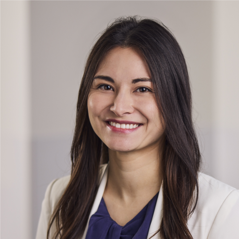 Femme aux longs cheveux foncés, vêtue d'un blazer blanc et d'un chemisier bleu marine, souriant à l'appareil photo à l'intérieur.