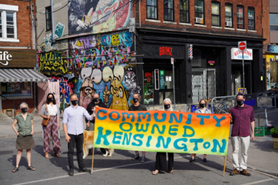Nine people hold a Community Owned Kensington banner, supporting affordable housing amid murals and shops.