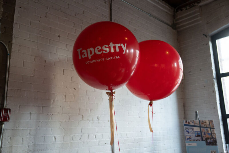 Deux grands ballons rouges portant l'inscription « Tapestry Community Capital », posés devant un mur de briques blanches à l'intérieur.