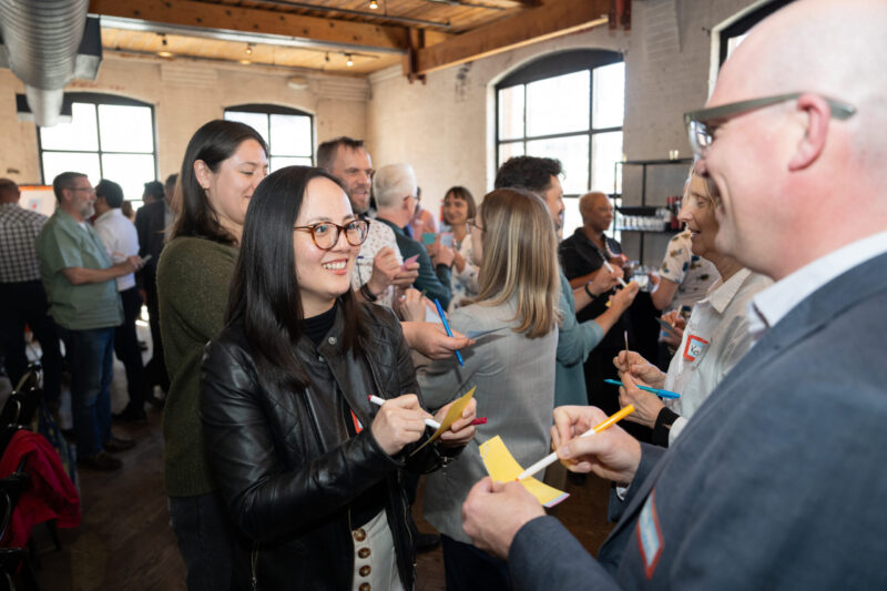 People standing in pairs, smiling and writing on sticky notes in a bright, busy room with large windows.