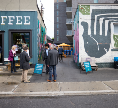 People stand near a coffee shop and colorful mural, with signs and outdoor seating visible in the alley.