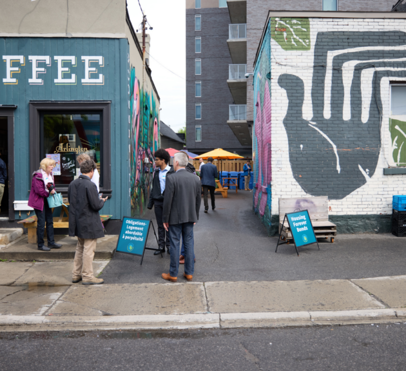 People stand near a coffee shop and colorful mural, with signs and outdoor seating visible in the alley.