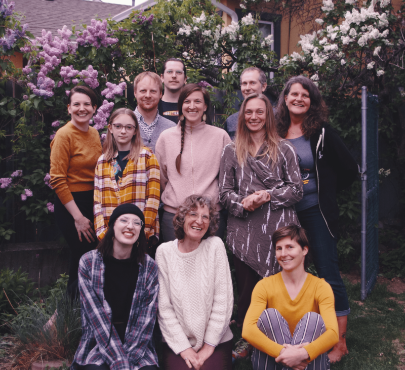 A group of eleven adults posing and smiling outdoors in a garden with lilac bushes in the background.