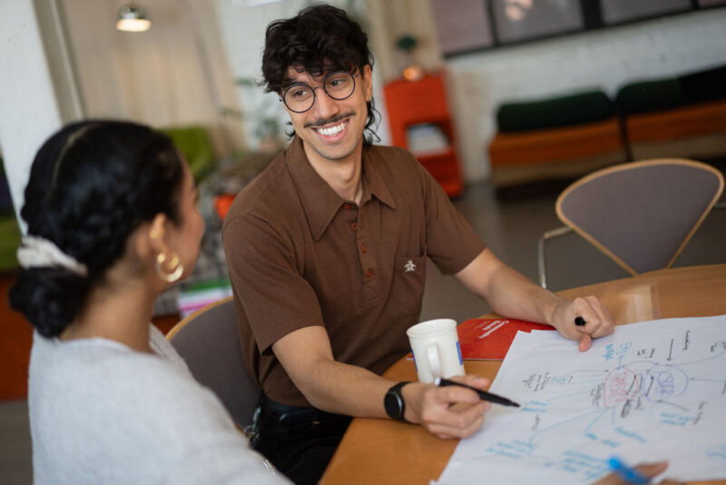 Deux personnes sont assises à une table, en train de discuter d'idées, le sourire aux lèvres, avec des notes et des schémas sur de grandes feuilles de papier devant elles.
