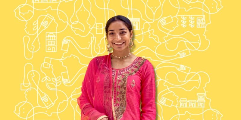 Une femme souriante, vêtue d'un costume traditionnel rose, se tient debout devant un fond jaune orné de motifs abstraits blancs dessinés au trait.