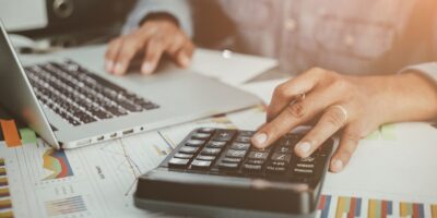 Hands using a calculator and laptop on a desk with charts and financial documents.