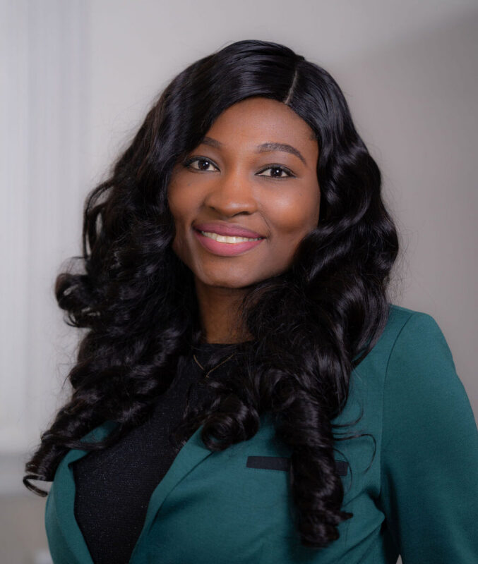 Woman with long curly hair in a green blazer, smiling at the camera against a neutral background.