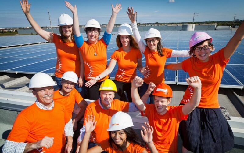 Un groupe de personnes vêtues de t-shirts orange et portant des casques de chantier font la fête sur un toit équipé de panneaux solaires.