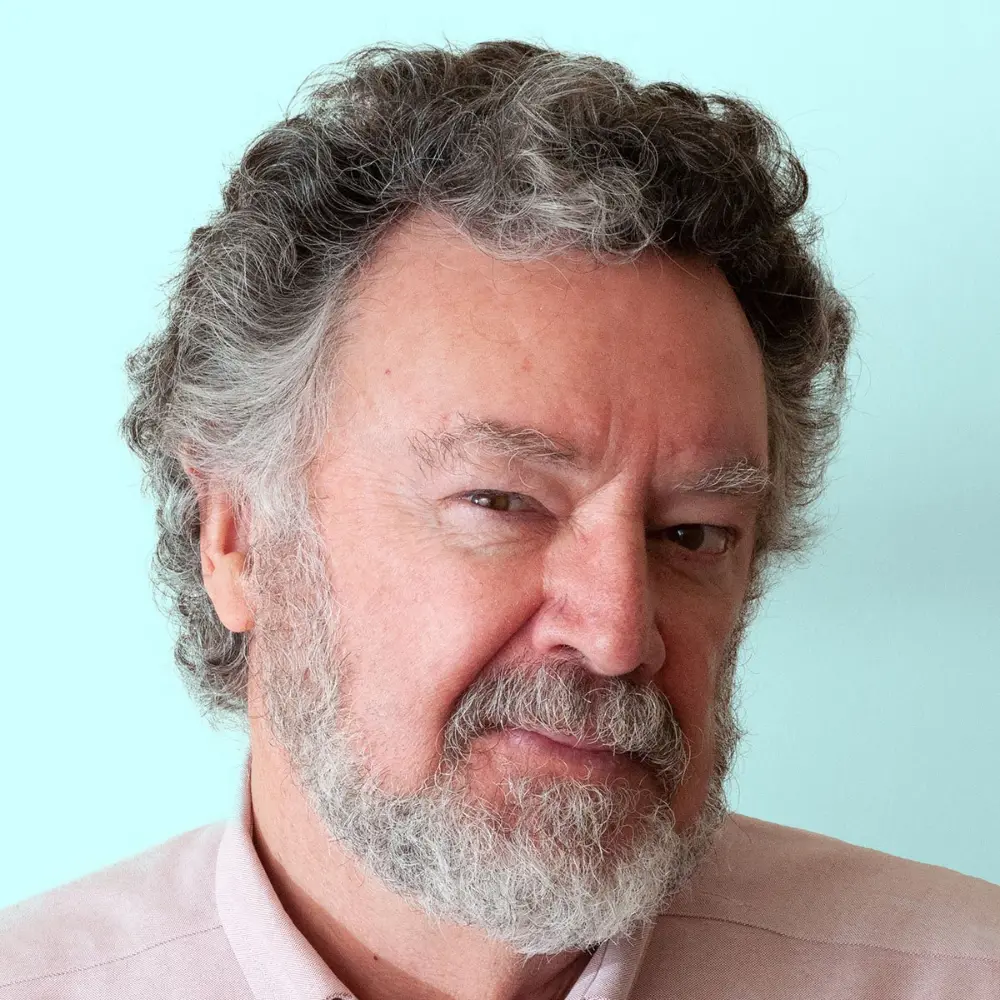 Older man with curly gray hair and beard, wearing a light pink shirt, looking seriously at the camera.
