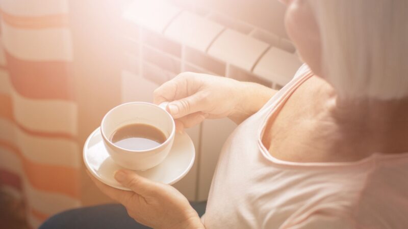 An older woman in soft sunlight holds a cup of coffee and saucer, sitting by a window with striped curtains.
