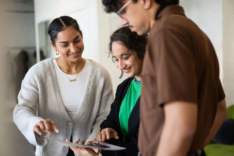 Three people standing together, looking at and pointing to a tablet, smiling and talking.