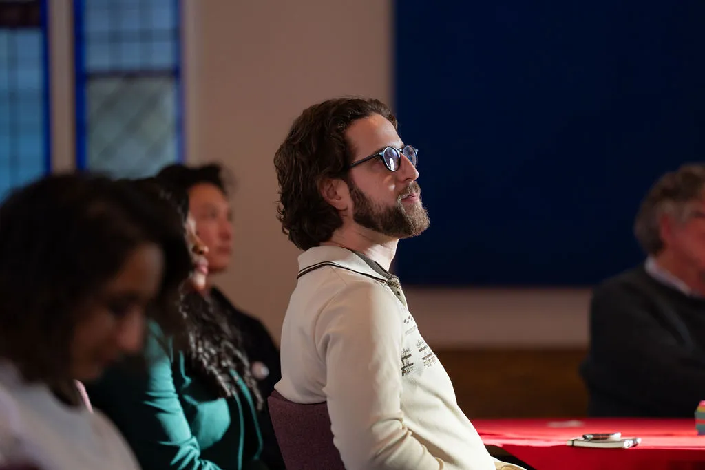 Un homme portant des lunettes et une barbe est assis, l'air attentif, parmi d'autres personnes dans une pièce baignée de soleil, le regard tourné vers l'avant.