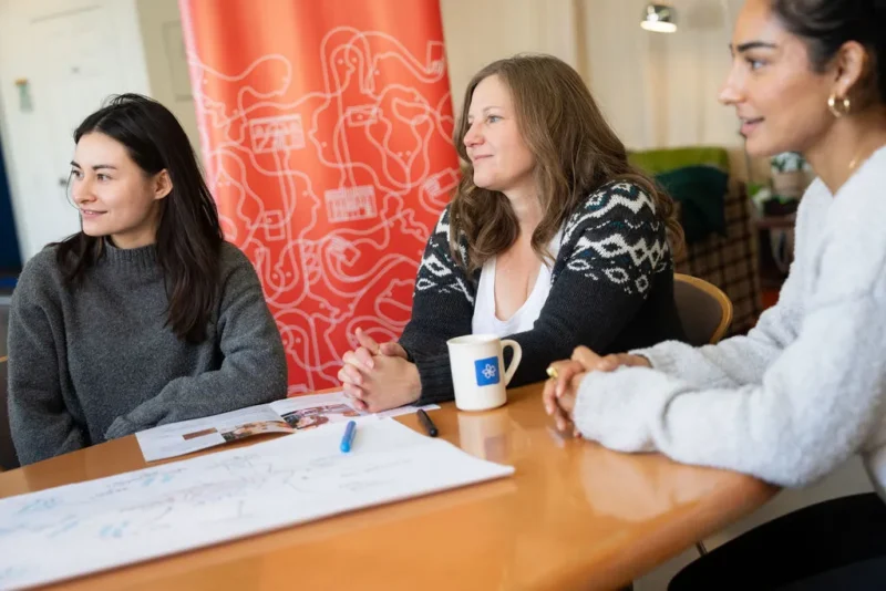 Three women sit at a table with papers and a pen, smiling and talking in a bright room.