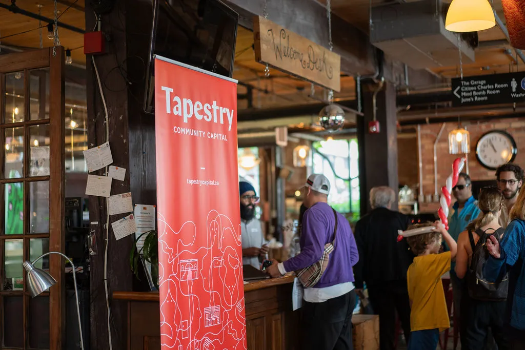 Indoor event with people gathered near a bar, next to a red Tapestry Community Capital banner.