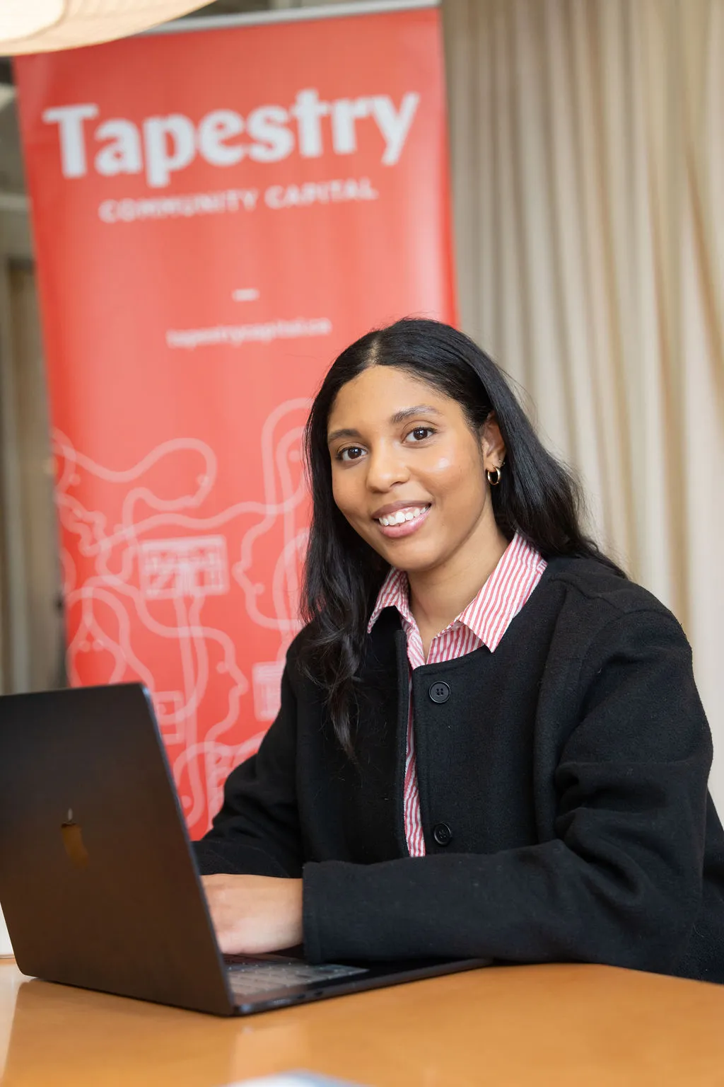 Woman smiling at a desk with a laptop, Tapestry Community Capital banner in background.