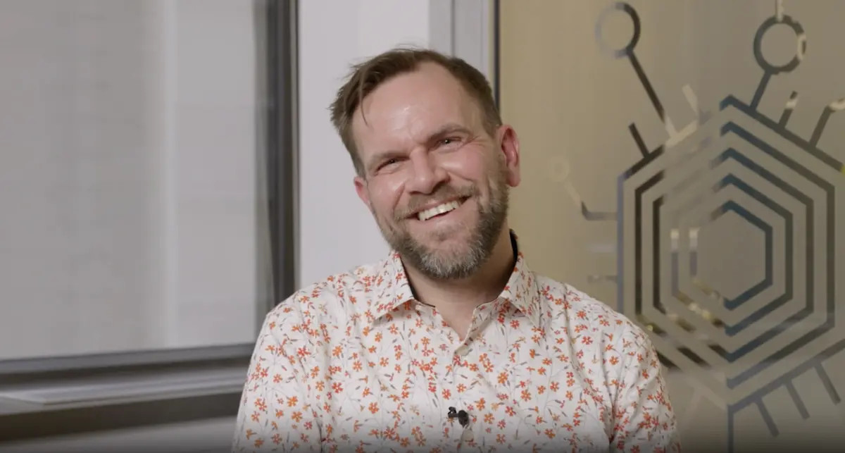 Un homme souriant, barbu, vêtu d'une chemise à fleurs, est assis à l'intérieur près d'une fenêtre et d'une décoration murale géométrique.