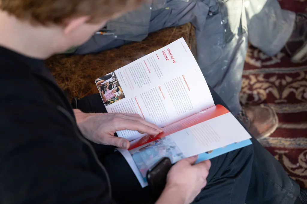 Une personne assise sur un tapis à motifs, en train de lire un magazine ou un livret ouvert contenant des images et du texte.