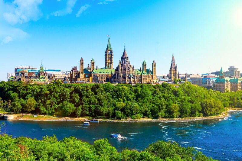 Ottawas Parliament buildings rise above green trees and a river under a bright blue sky.