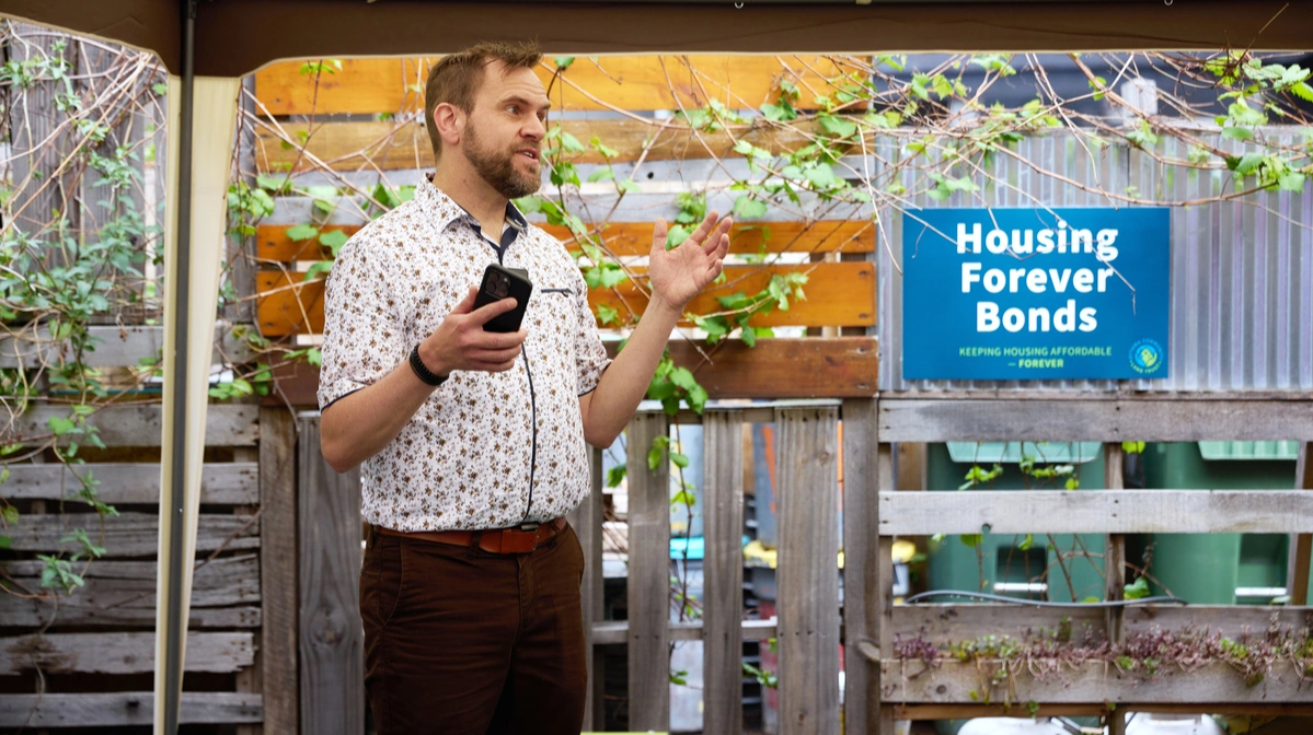 Un homme parlant en extérieur près d'un panneau « Housing Forever Bonds », un téléphone à la main, avec des plantes en arrière-plan.