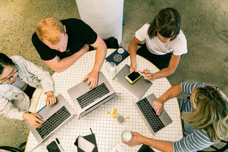 Four people work on laptops and a tablet at a round table with drinks, seen from above.