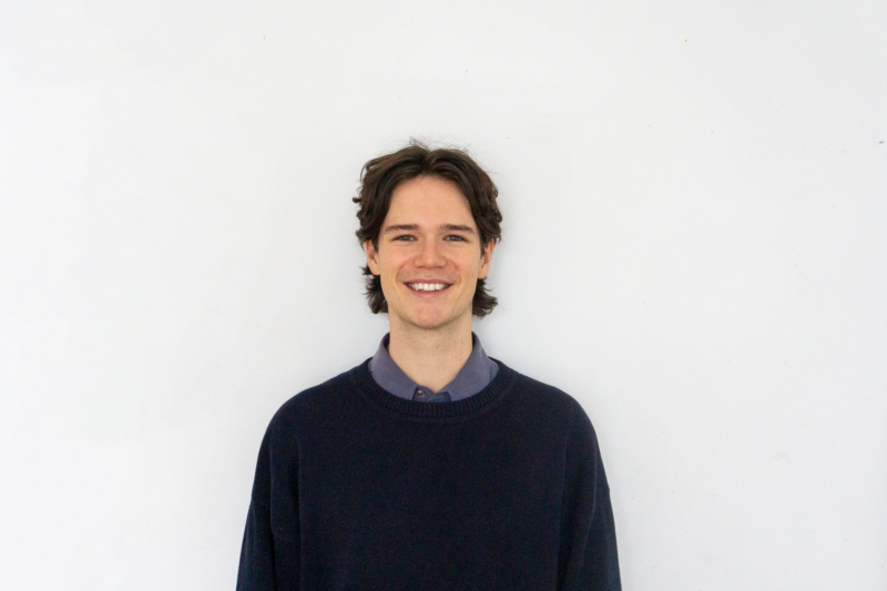 A young person with brown hair smiles at the camera, standing against a plain white background.
