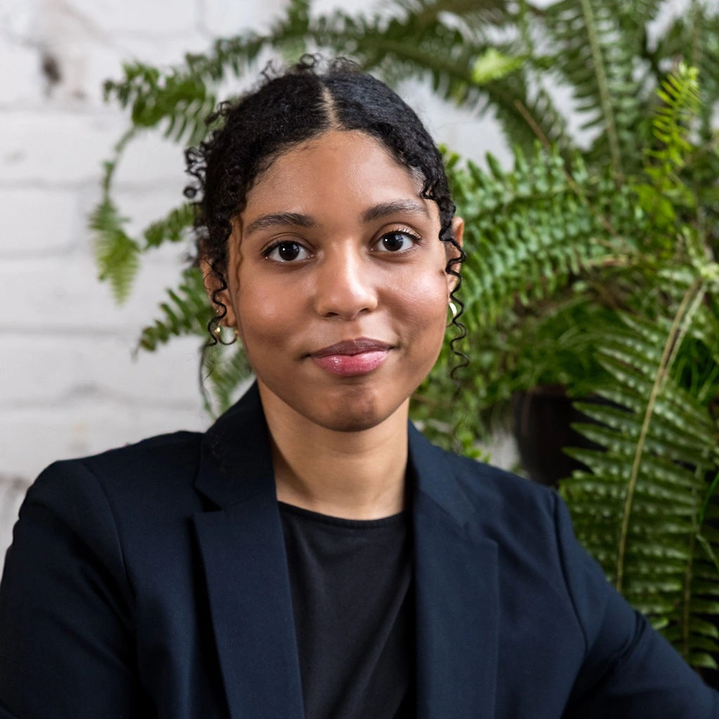 Woman with curly hair in a black blazer sits in front of green ferns and a white brick wall.