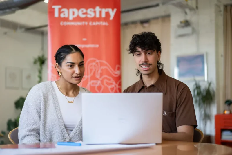 Deux personnes sont assises côte à côte et regardent l'écran d'un ordinateur portable. Une bannière rouge « Tapestry » est visible derrière elles.
