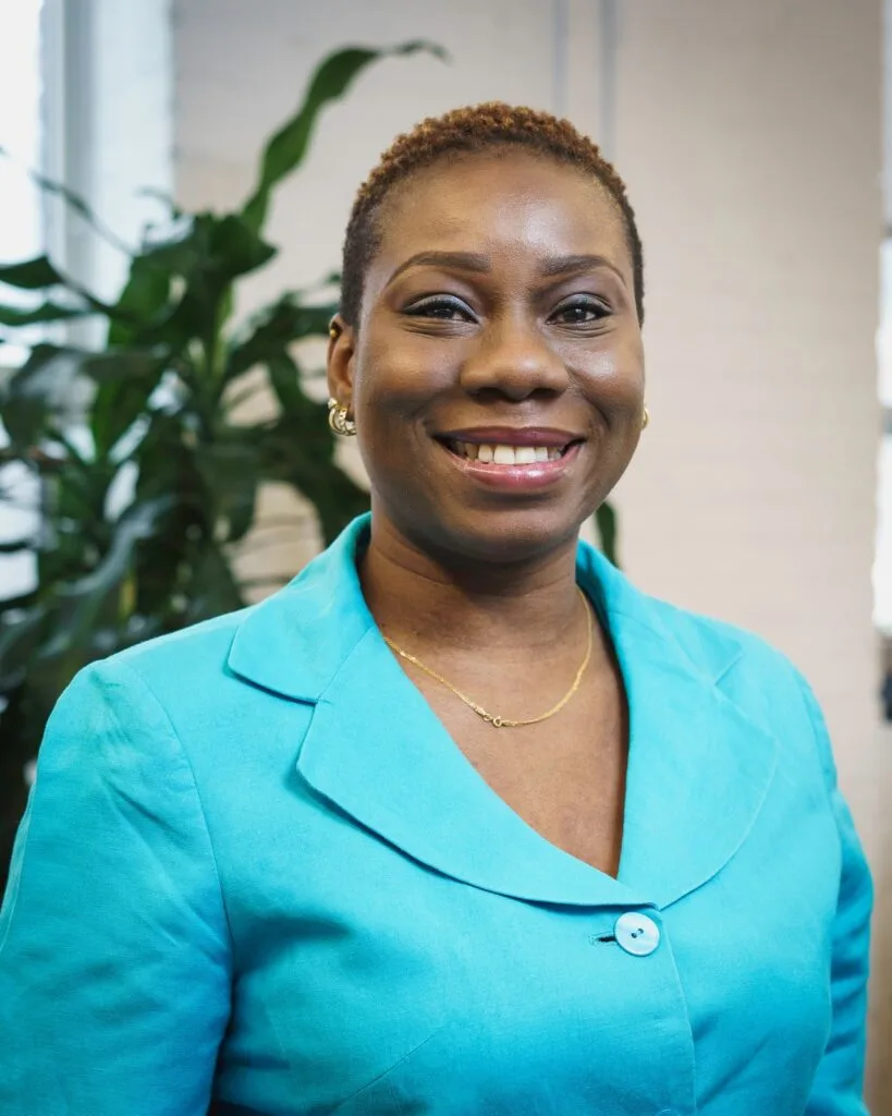 Smiling woman in a turquoise blazer stands indoors with a plant in the background.