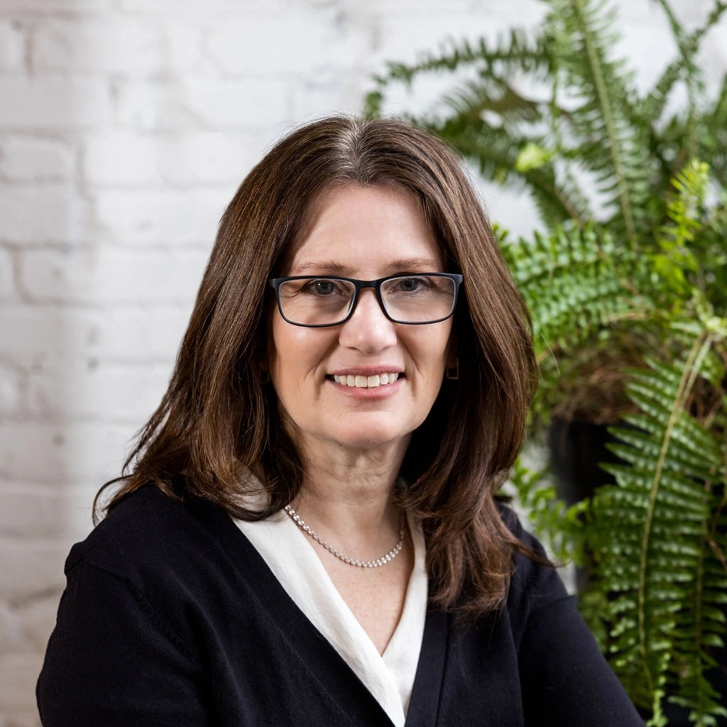 Woman with brown hair and glasses smiling, sitting in front of a white brick wall and green fern plant.