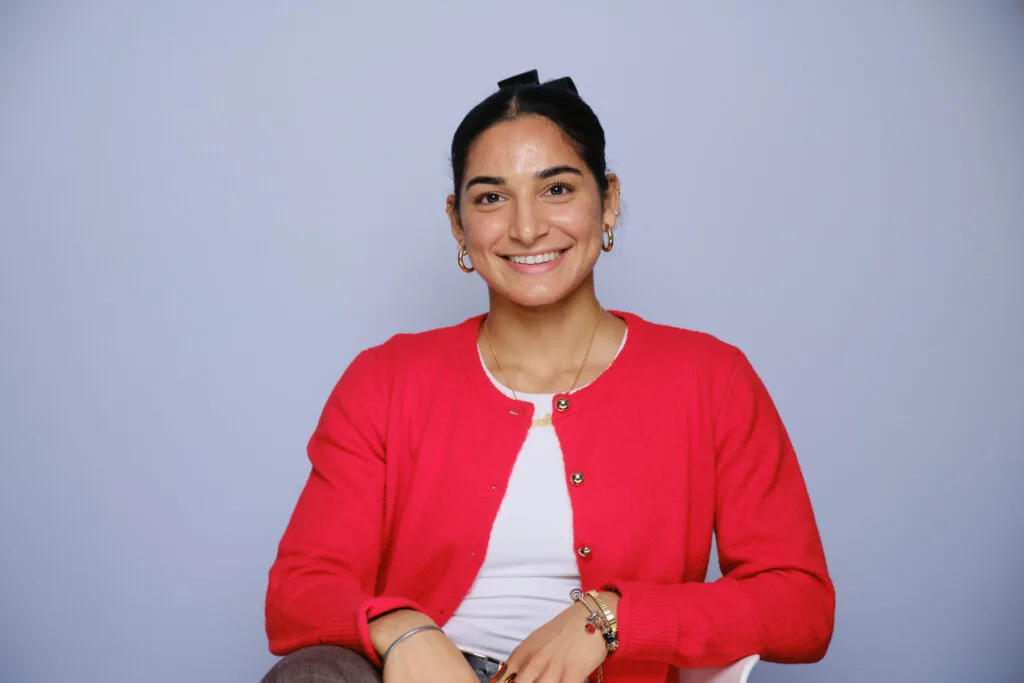 Woman with dark hair, wearing a red cardigan, white shirt, and bracelets, smiling against a plain background.