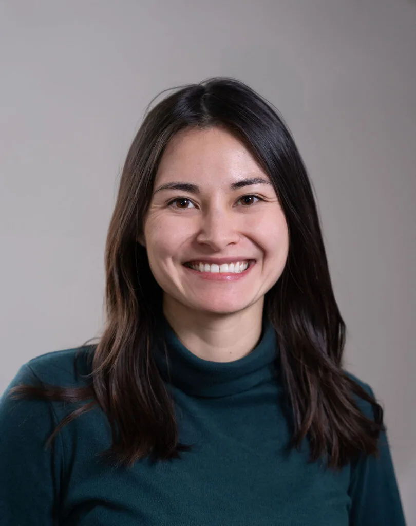 Woman with long dark hair wearing a dark green turtleneck, smiling at the camera against a gray background.