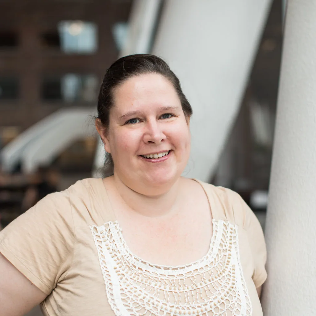 Woman smiling indoors, wearing a beige shirt with a white lace front, blurred background.