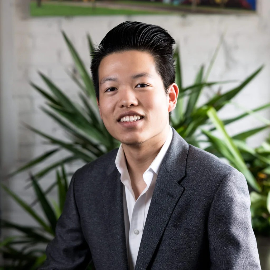 Man in a gray suit jacket and white shirt smiling, with green plants in the background.