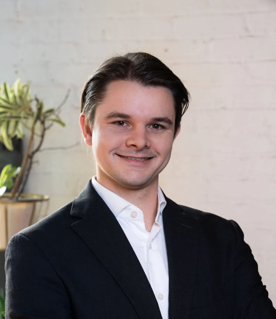 Man in a dark suit and white shirt smiling, standing in front of a white brick wall with a plant.